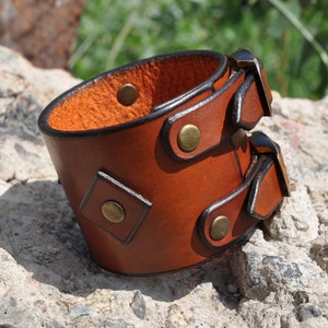 May include: A wide brown leather cuff bracelet featuring two adjustable buckle straps with brass-toned metal hardware. The exterior surface is smooth, while the interior shows a textured, natural leather finish with visible brass rivets.