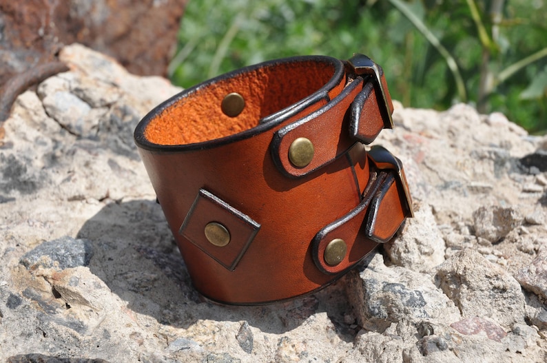 May include: A wide brown leather cuff bracelet featuring two adjustable buckle straps with brass-toned metal hardware. The exterior surface is smooth, while the interior shows a textured, natural leather finish with visible brass rivets.