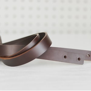 May include: Two brown leather straps with holes for stitching. The straps are twisted together and are lying on a white surface.