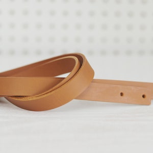 May include: Two pieces of brown leather strap with holes for stitching. The straps are folded and lying on a white surface.