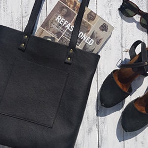 Könnte beinhalten: Eine schwarze Ledertasche mit einem vorderen Fach und goldfarbenen Beschlägen. Die Tasche liegt auf einer weißen Holzoberfläche mit einem Buch und einer Sonnenbrille.