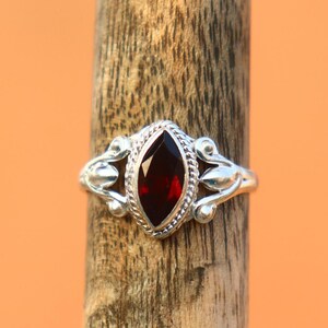 May include: A silver ring with a marquise-cut, deep red gemstone. The ring features ornate detailing on the band and around the stone. The ring is displayed on a wooden dowel against an orange background. Jewelry.