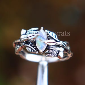 May include: A silver ring with a leaf and vine design, featuring a central, oval-shaped, iridescent gemstone. The ring's band and setting are intricately detailed with leaves and delicate metalwork, creating a nature-inspired aesthetic.