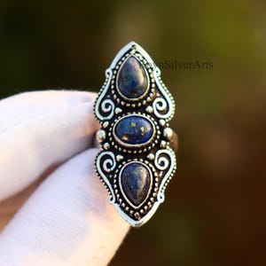 May include: A silver-toned ring featuring three dark blue gemstones. The ring has an elongated design with ornate scrollwork and a decorative setting around the stones. The gemstones are oval and teardrop-shaped.