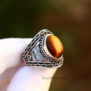 May include: A silver ring featuring an oval, amber-hued tiger's eye gemstone. The ring has an intricate, filigree pattern around the stone and on the band. The ring is shown against a blurred backdrop.
