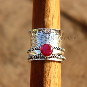 May include: A silver-colored spinner ring with a round, red gemstone at the center. The ring has a floral pattern on the top band and a twisted rope design on the bands below and above the gemstone. The ring is displayed on a wooden dowel.
