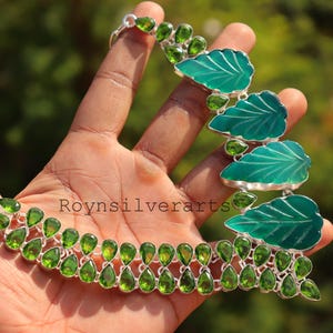 Può includere: Una collana d'argento con pietre preziose verdi e ciondoli a forma di foglia. La collana presenta pietre verdi a forma di goccia e ciondoli più grandi a forma di foglia con finitura in smalto verde acqua. La collana è tenuta in una mano.