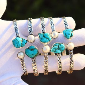 Peut inclure: Plusieurs bracelets en argent, chacun orné d'une pierre turquoise unique et d'une perle. Les bracelets présentent un motif gravé ornemental sur le bracelet. Les pierres turquoises varient en forme et en taille, mettant en valeur les textures naturelles.