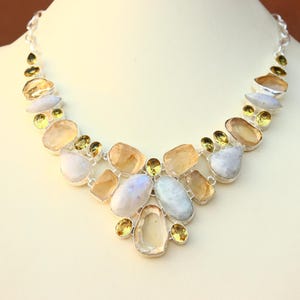 May include: A silver necklace featuring a variety of gemstones in shades of yellow, white, and blue. The necklace is displayed on a mannequin, showcasing its design and color palette. The gemstones are of various shapes and sizes.