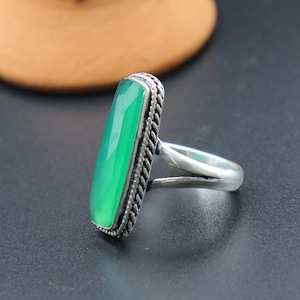 Może przedstawiać: Srebrny pierścionek z dużym, prostokątnym, fasetowanym zielonym kamieniem szlachetnym. Kamień osadzony jest w ozdobnej, sznurowatej obwódce. Pierścionek jest prezentowany na ciemnej powierzchni, z rozmytym tłem.