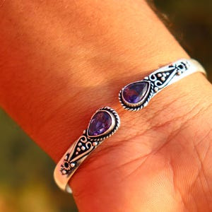 Puede incluir: Brazalete de plata con detalles ornamentados y dos gemas moradas en forma de lágrima. El diseño abierto facilita su uso. El brazalete se muestra en una muñeca.