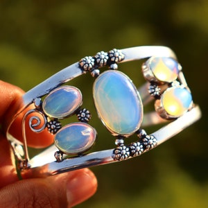 Puede incluir: Un brazalete de plata con un diseño delicado que presenta múltiples piedras de ópalo iridiscentes. El brazalete tiene una forma curva y un motivo floral.
