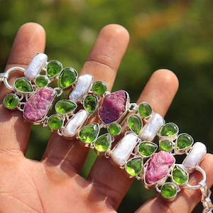 May include: A silver bracelet featuring a series of gemstones. The bracelet incorporates large, rough-cut pink stones, interspersed with green, pear-shaped stones and white, oblong pearls. The bracelet is held in a hand against a blurred green background.