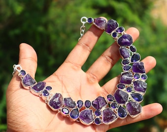 May include: A handcrafted necklace featuring large, irregular amethyst stones and smaller, polished purple gems set in a silver-tone metal. The necklace is displayed against a blurred green background.