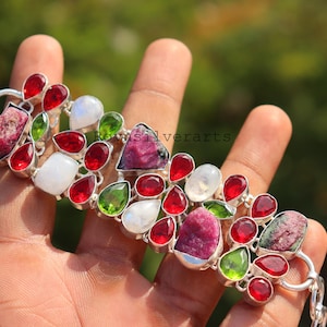 May include: A silver bracelet featuring a cluster of colorful gemstones. The bracelet showcases a variety of stones in shades of red, green, white, and pink. The gemstones are set in a silver metal setting.