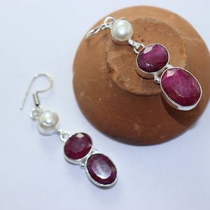 Puede incluir: Un par de pendientes de plata con una perla blanca y dos piedras de rubí rojo. Los pendientes son colgantes y tienen un cierre de gancho.