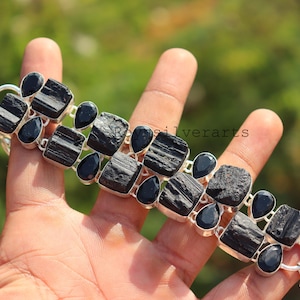 May include: A silver bracelet featuring large, rough-cut black tourmaline stones and smaller, teardrop-shaped black onyx stones. The bracelet is displayed against a blurred green background, showcasing the natural beauty of the gemstones.