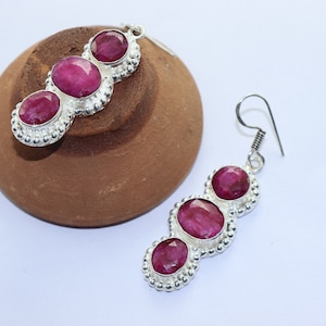Puede incluir: Pendientes de plata con tres piedras preciosas ovaladas de color rojo intenso. Cada gema está engastada en un marco de plata con un borde de cuentas. Se muestran sobre un fondo blanco.