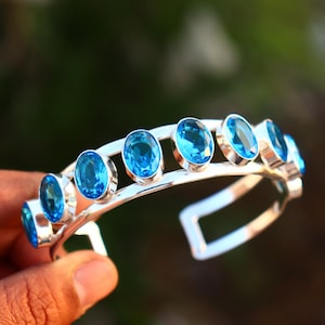 Pode incluir: Uma pulseira de punho prateada com pedras preciosas ovais azuis brilhantes. As pedras preciosas são colocadas em biséis individuais ao longo da parte superior da pulseira. A pulseira é segurada em uma mão contra um fundo desfocado verde e marrom.