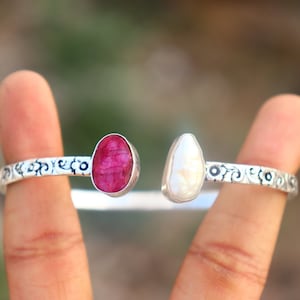 May include: A silver bangle bracelet with an ornate design features a red ruby and a white pearl.