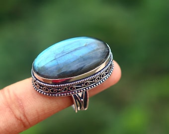 May include: An oval-shaped labradorite gemstone ring featuring a polished, iridescent blue and grey stone set in a silver band. The metal setting displays intricate, embossed scrollwork patterns along the sides and a beaded edge detail.