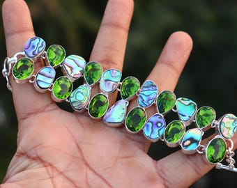 May include: A silver bracelet featuring a series of oval-shaped green gemstones and iridescent abalone shell accents. The bracelet is displayed against a blurred background, showcasing the jewelry's vibrant colors and intricate design.