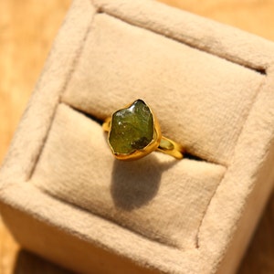 May include: A gold ring with a large, green, rough-cut gemstone. The ring is in a small, beige velvet box.