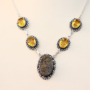 Könnte beinhalten: Eine silberne Halskette mit einem zentralen ovalen Stein und vier gelben ovalen Steinen, alle in dekorativen Rahmen gefasst. Die Halskette ist durch silberne Glieder verbunden. Das Design wird vor einem hellen Hintergrund präsentiert. Schmuck.