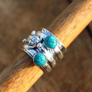 Può includere: Un anello d'argento con un design a tartaruga e due pietre turchesi. L'anello ha una fascia strutturata ed è posto su uno sfondo di legno. L'anello è un gioiello.