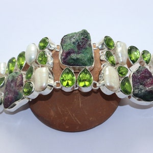以下が含まれることがあります： 緑とピンクの宝石と白い真珠が付いたシルバーブレスレット。ブレスレットは留め金で留まります。