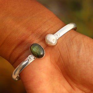 Puede incluir: Pulsera de brazalete de plata con una piedra preciosa de labradorita verde y una perla blanca. La pulsera es abierta y ajustable.