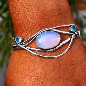 Puede incluir: Una pulsera de plata con una piedra central ovalada y opalescente y dos pequeñas piedras azules redondas. La pulsera tiene un diseño fluido y orgánico. La joya se muestra en un brazo.