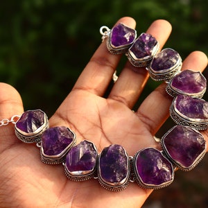 Puede incluir: Un collar de plata con grandes piedras de amatista facetadas. Las piedras están engastadas en engastes de plata y están conectadas por una cadena de plata.