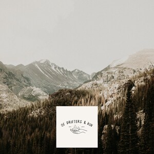 May include: A white square with the text "OF DRIFTERS & KIN" in black lettering. The text is surrounded by a simple line drawing of a hand holding a mountain range.