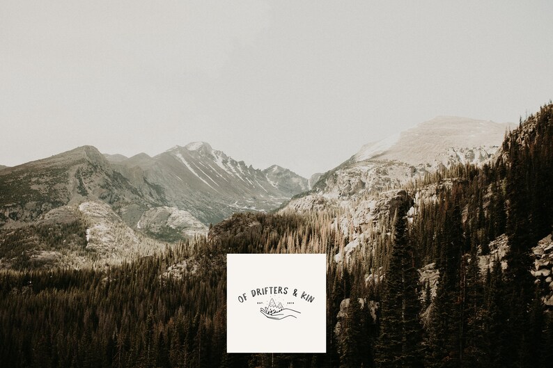 May include: A white square with the text "OF DRIFTERS & KIN" in black lettering. The text is surrounded by a simple line drawing of a hand holding a mountain range.