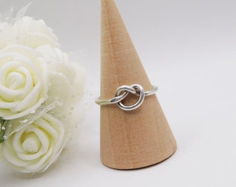 Op de afbeelding: Een zilveren knoop ring op een houten ringstandaard. De ring is een eenvoudige band met een knoopontwerp in het midden.