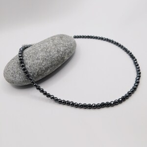 May include: A black hematite beaded necklace with a silver clasp. The necklace is lying on a gray rock.
