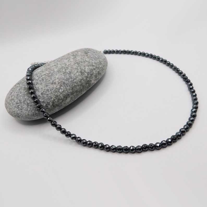 May include: A black hematite beaded necklace with a silver clasp. The necklace is lying on a gray rock.