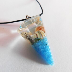 Könnte beinhalten: Ein klarer Harz-Anhänger in Pyramidenform mit einer schwarzen Schnur. Der Anhänger enthält Muscheln und blauen Sand.