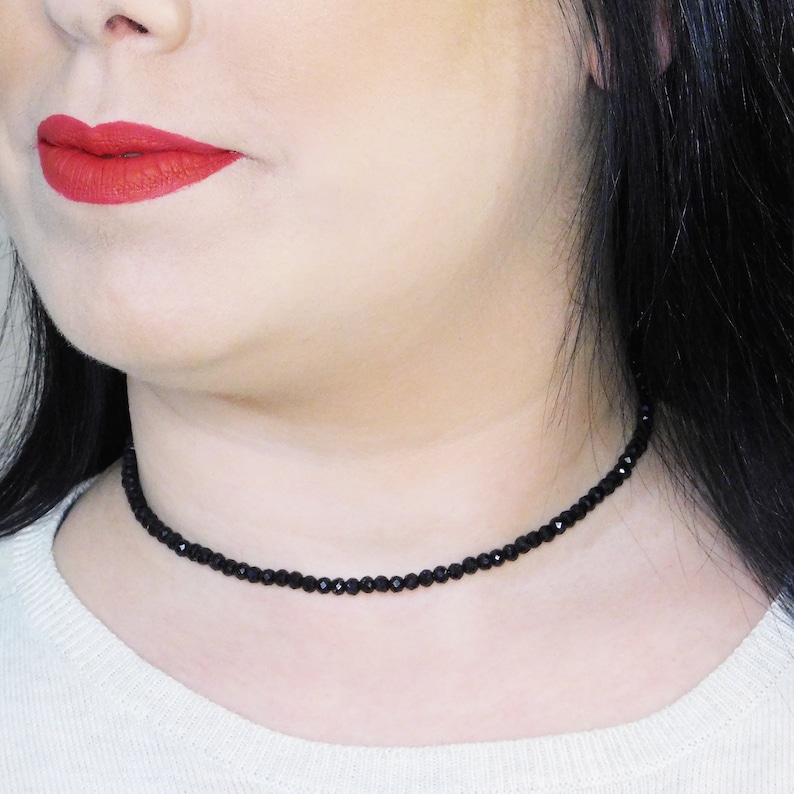 May include: A black choker necklace made of small, faceted beads. The necklace is worn by a person with dark hair and red lipstick.