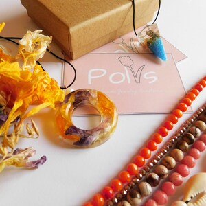 May include: Two resin necklaces with dried flowers encased in the resin. One necklace has a round pendant with a hole in the center. The other necklace has a teardrop pendant with blue and white resin. The necklaces are displayed on a white surface with a business card that says "Polvs Handmade Jewelry Creations".