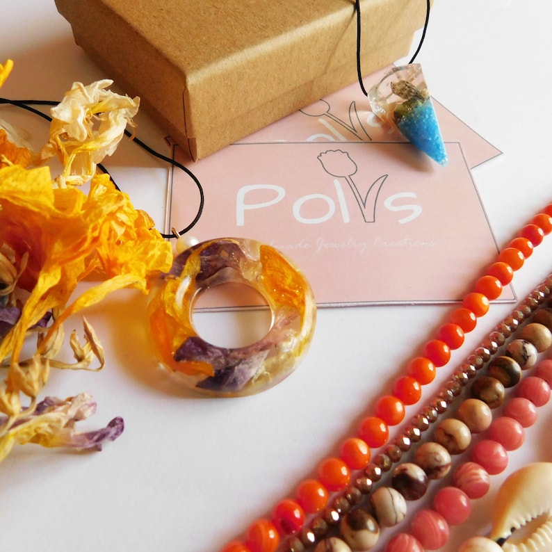 May include: Two resin necklaces with dried flowers encased in the resin. One necklace has a round pendant with a hole in the center. The other necklace has a teardrop pendant with blue and white resin. The necklaces are displayed on a white surface with a business card that says "Polvs Handmade Jewelry Creations".