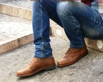 May include: A pair of brown leather ankle boots with laces. The boots are worn by a person wearing blue jeans and a red and white plaid shirt.