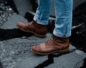 May include: A pair of brown leather boots with dark brown laces and a cap toe. The boots are worn with blue jeans with the cuffs rolled up. The boots are on a cracked, dark gray surface.