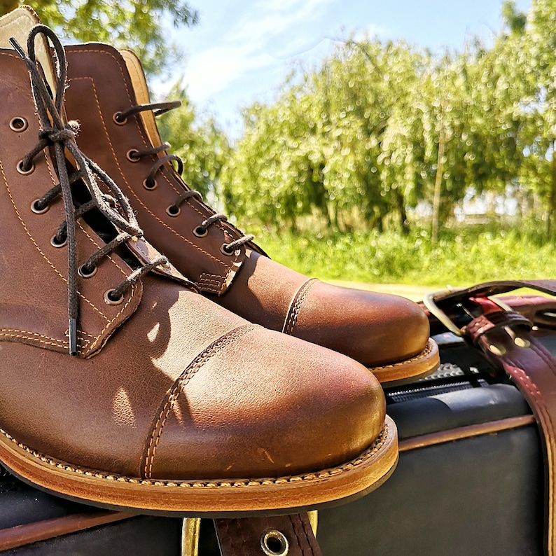 May include: A pair of brown leather boots with laces. The boots have a cap toe design and are sitting on a brown leather surface.