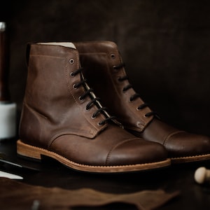 May include: A pair of brown leather lace-up boots with a cap toe design. The boots are on a dark surface with leather working tools.