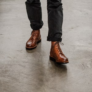 Puede incluir: Un par de botas de cuero marr&oacute;n con cordones y punta ligeramente puntiaguda. Las botas las lleva una persona que lleva pantalones oscuros.