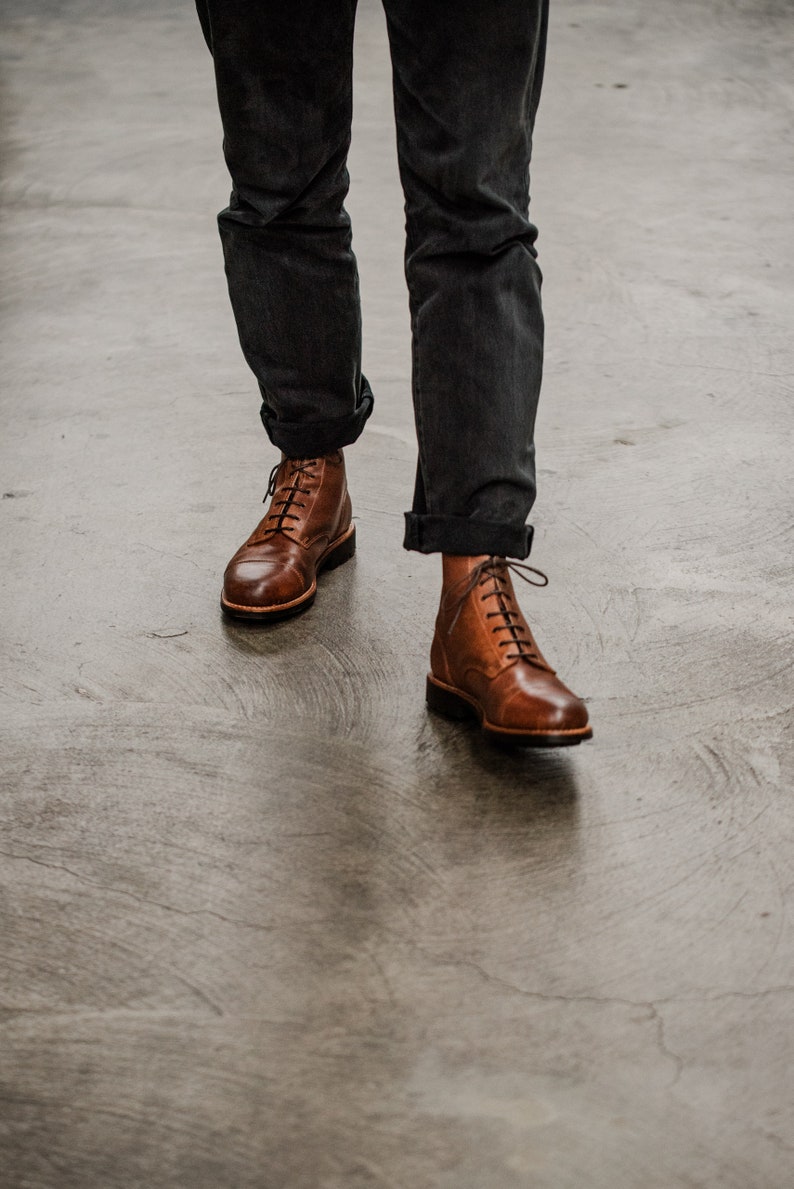 Puede incluir: Un par de botas de cuero marr&oacute;n con cordones y punta ligeramente puntiaguda. Las botas las lleva una persona que lleva pantalones oscuros.