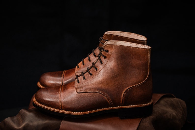 Puede incluir: Un par de botines de cuero marr&oacute;n con cordones. Las botas tienen un aspecto ligeramente desgastado y est&aacute;n sobre una superficie de cuero marr&oacute;n.