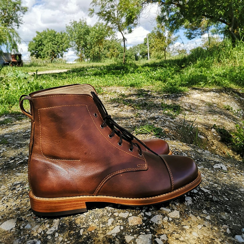 May include: A pair of brown leather lace-up boots with a cap toe design. The boots are sitting on a rocky surface with a green grassy background.